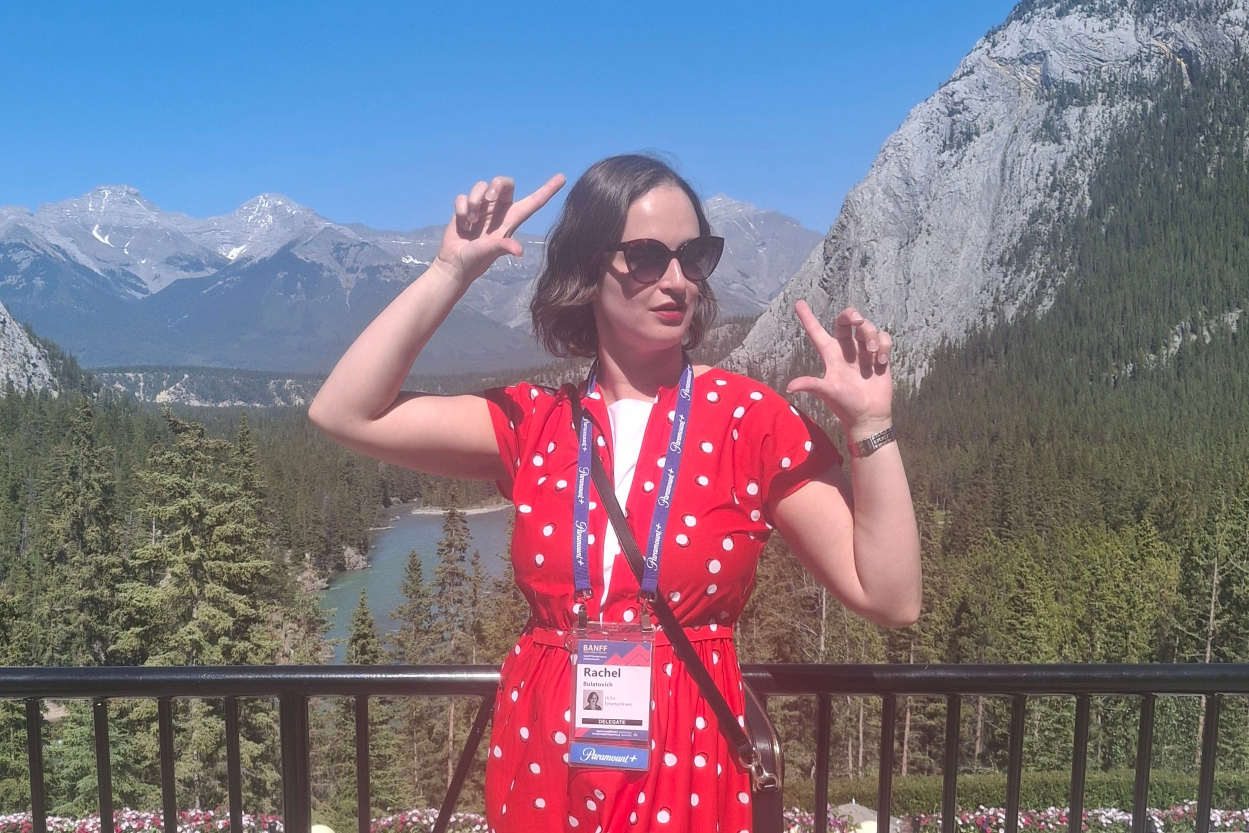 A photo of Rachel Bulatovich - she has chin-length brown hair and wears brown sunglasses and a red and white polka dot dress. She stands in front of a mountain range framing her face with her fingers.